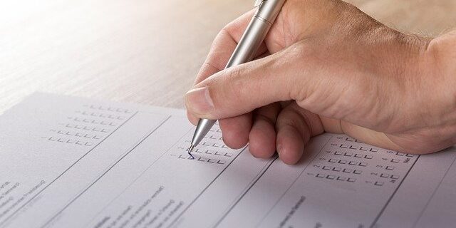 Hand holding pen, filling out a tick-box form or survey on a sheet of paper on a desk.
