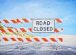 A Road Closed sign blocks a deserted road with sand dunes and blue sky in the background.