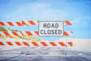 A Road Closed sign blocks a deserted road with sand dunes and blue sky in the background.
