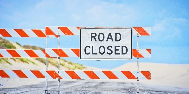 A Road Closed sign blocks a deserted road with sand dunes and blue sky in the background.