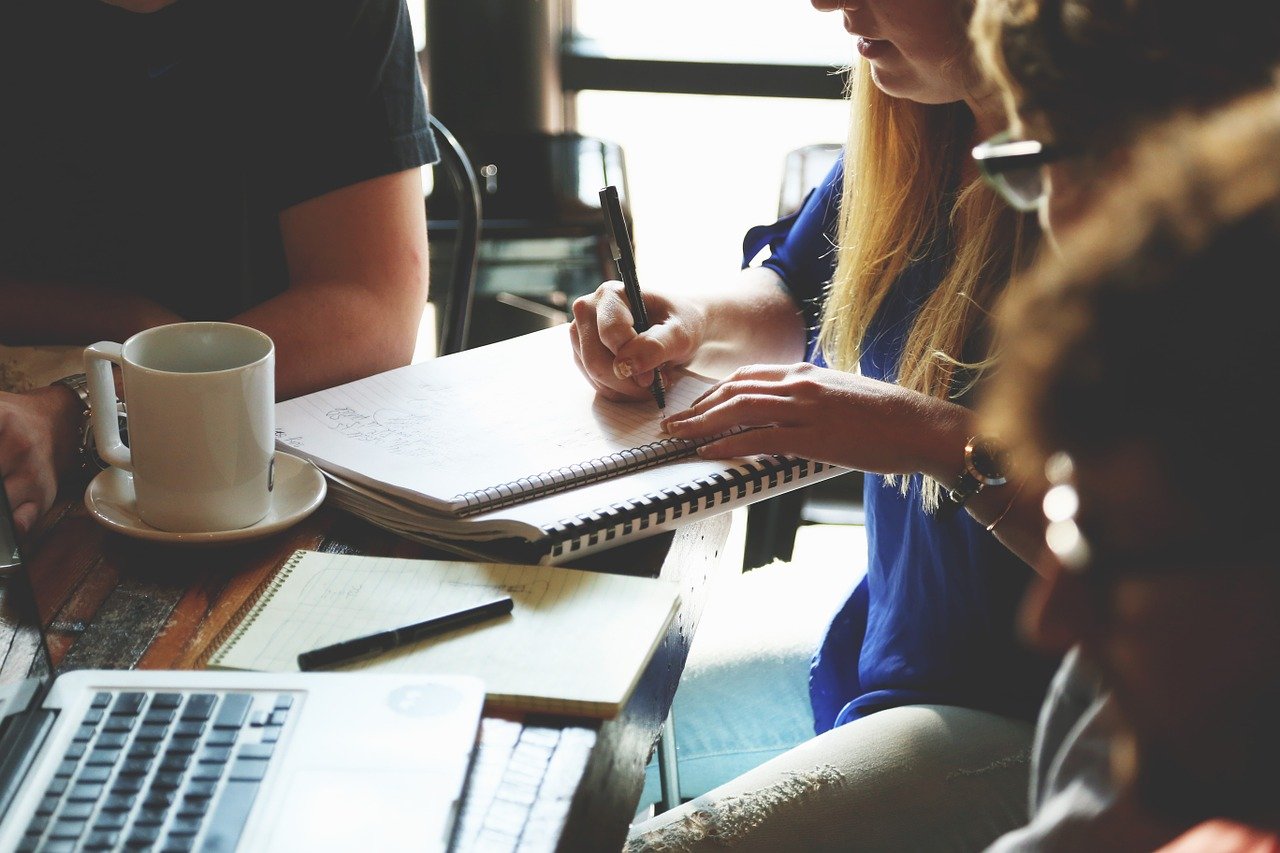 People sitting at a table, writing in notebooks with coffee and a laptop nearby.