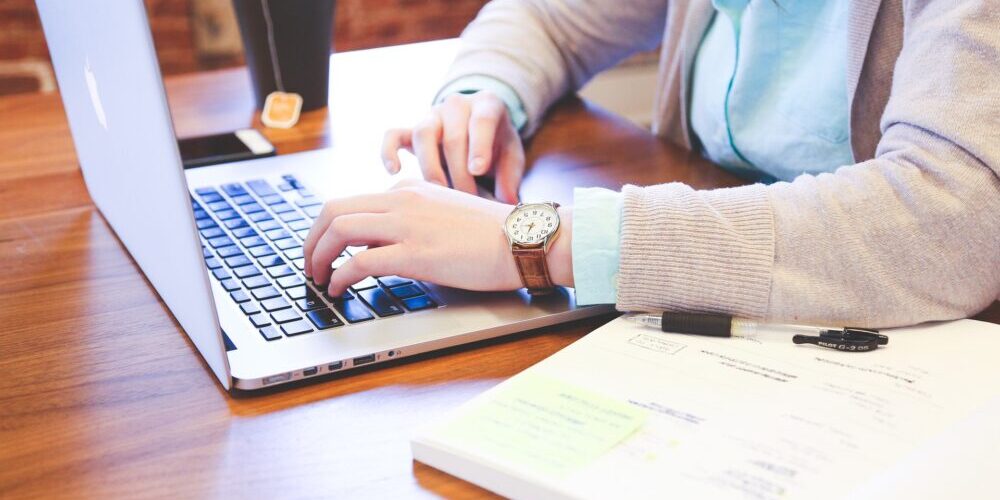 Person typing on a laptop at a wooden desk with papers, a pen, and a cup of tea nearby.