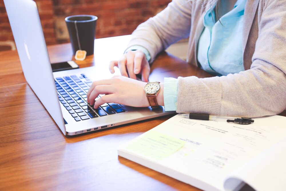 Person typing on a laptop at a wooden desk with papers, a pen, and a cup of tea nearby.