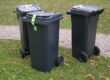Three black wheelie bins with green labels on grass near a paved path in a park.