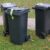 Three black wheelie bins with green labels on grass near a paved path in a park.