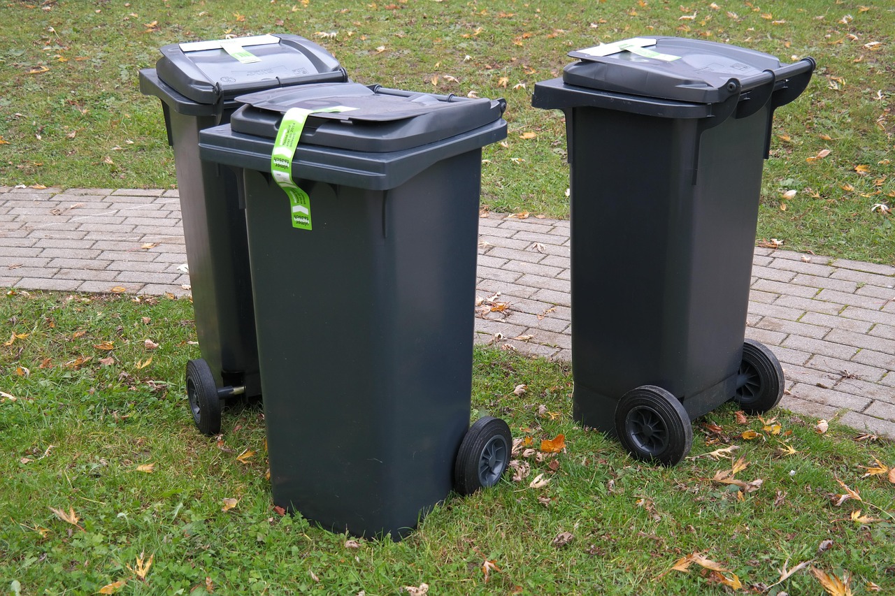 Three black wheelie bins with green labels on grass near a paved path in a park.